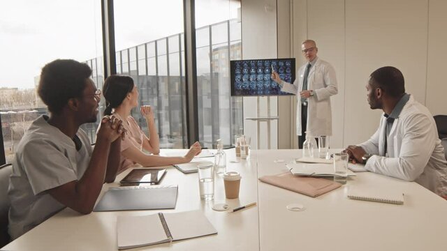 Medium slowmo of mature radiologist or neurologist in lab coat standing next to display monitor with brain x-ray image talking to young multiethnic medical interns or colleagues