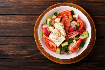 Radish, cucumber, tomato, pepper and feta cheese with spices pepper and olive oil in white bowl on old wooden dark table background. Healthy food concept. Top view.