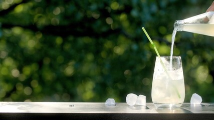 Making drinks outdoors. Pouring carbonated drink into a transparent glass with ice. Straw in a drink. Many ice cubes in a glass and around. 