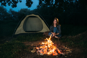 smiling lonely girl tourist in the forest traveler sitting by the fire near the tent in the camping in the evening after sunset. Autumn evening and a young woman is resting by the fire.