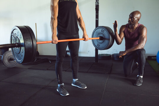 Diverse fitness trainer and man exercising at gym, lifting weights on barbell and advising