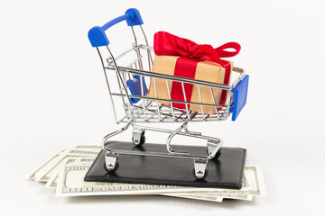 metal chrome shiny shopping trolley on wheels with gift box on wallet with dollars on white background. selective focus