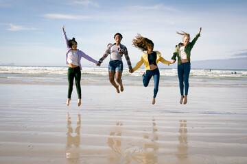 Happy group of diverse female friends having fun, walking along beach holding hands and jumping