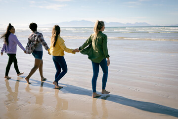 Happy group of diverse female friends having fun, walking along beach holding hands and laughing