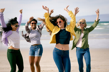 Happy group of diverse female friends having fun, walking along beach holding hands and laughing