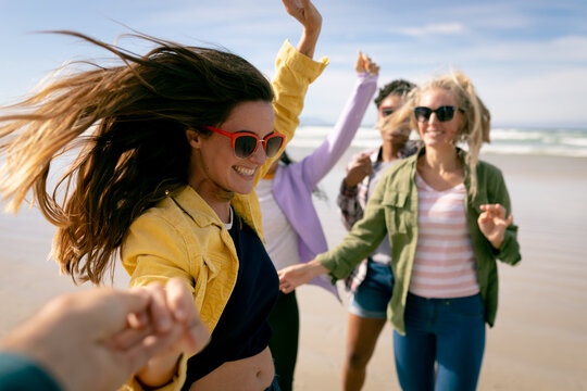 Happy group of diverse female friends having fun, walking along beach holding hands and laughing