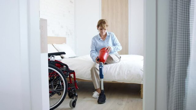 Disabled mature woman sitting on bed indoors at home, morning routine with leg amputee concept.