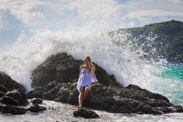 A young girl with long hair sits on a stone by the sea, from behind the wave breaks on a stone with...
