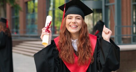 Educational concept. Happy caucasian student girl being happy at her diploma, graduation day. Ginger woman standing at the street with her friends at the background