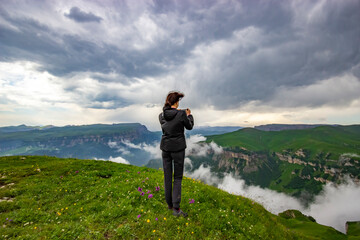 A girl stands on the edge of a cliff and takes pictures of a beautiful view. View from the back. Rest in the mountains. The concept of active recreation. Tourism in the highlands. Rest in solitude.