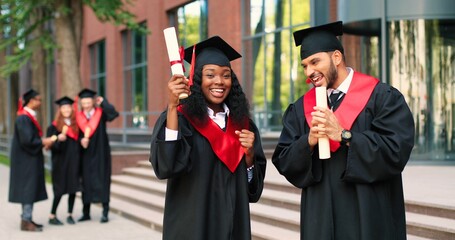 Two happy multiracial student people are celebrating their graduation while standing near the...