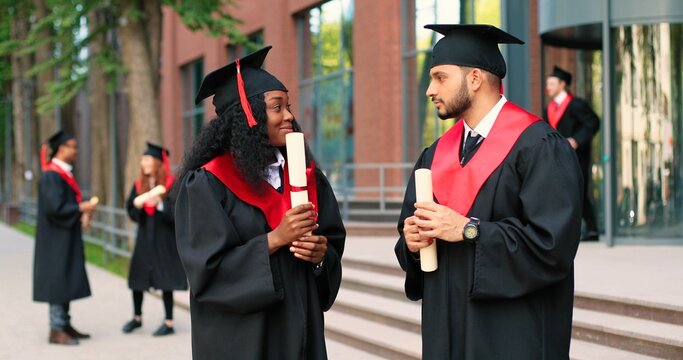 Two happy multiracial student people are celebrating their graduation while standing near the college and rejoicing with diplomas at the hands. Education concept
