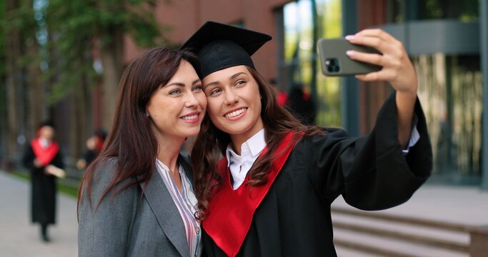 Happy mother and her graduate daughter posing for selfie on graduation day. Girl in academic dress and caps taking self picture with her proud mother