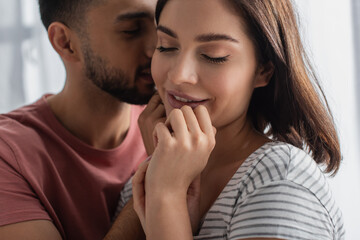 young man kissing smiling girlfriend with closed eyes and hands near face in kitchen