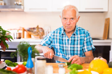 elderly man cuts vegetables for salad at the table in the kitchen