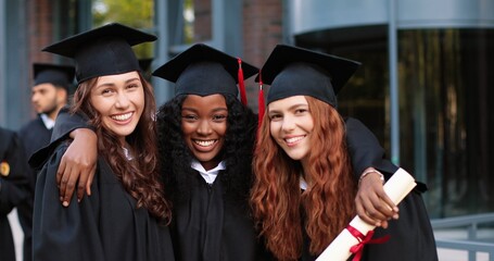 Fototapeta Happy group of mature students on graduation day embracing with each other. Three best girl friends in academic gowns and caps hugging in front of the camera obraz
