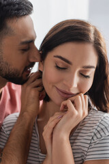 young man kissing smiling woman with closed eyes and hands near face at home