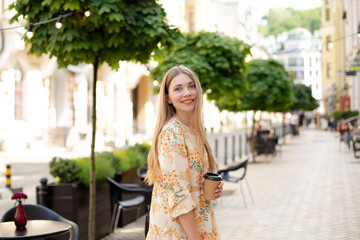 caucasian smiling blonde woman in a dress drinking coffee takeaway and walking down the street against the background of green trees and yellow lanterns, close-up