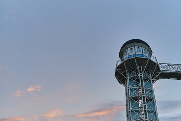 Minimalist landscape at dawn and high elevator to sky with bridge. Pink beautiful natural clouds in clear blue morning sky. Unusual lighthouse with Windows view from below.
