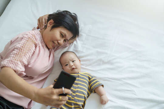 Happy Asian Grandmother and little baby boy or Grandson lying on bed taking selfie photos and doing video call with mobile phone or smartphone.