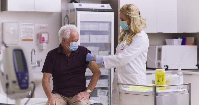 A Wide Shot Of An American Female Medical Doctor Administering A Covid-19, SARS-CoV-2 Vaccine Injection With A Needle and Syringe, To An Elderly Man. Wearing Safety Gloves And Protective Mask
