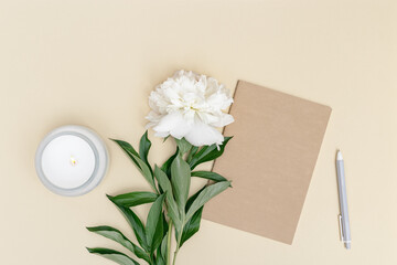 Feminine desktop, closed notebook, pen, fresh white peony flower and lit fragrant candle in glass jar on beige. Summer work concept.