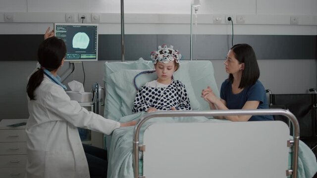 Pediatrician woman doctor discussing tomography expertise while monitoring sickness evolution during recovery consultation. Sick child wearing eeg brain sensors headset in hospital ward