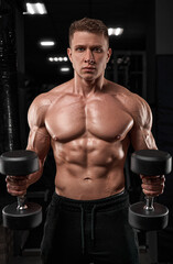 Muscular bodybuilder doing exercises with dumbbell in gym.Strong athletic man shows body, abdominal muscles, biceps and triceps.Work out, gaining weight, pumping up muscles with dumbbells.