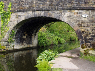 Fototapeta na wymiar an old stone bridge crossing the rochdale canal at mytholmroyd west yorkshire with a waterside pathway surrounded by trees and bushes