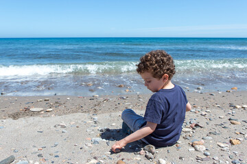 Curly-haired boy with Caucasian appearance, sitting by the sea, picking up stones from the beach