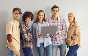 Portrait of happy IT students with laptop computer smiling and looking at camera standing together on gray studio background. Group of young people working on project and using online learning tools - obrazy, fototapety, plakaty
