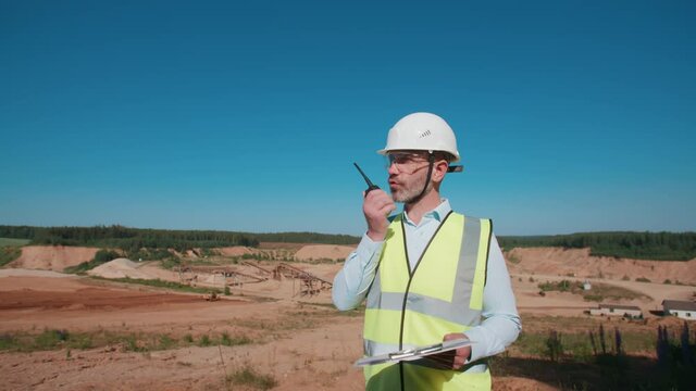 Handsome male sand quarry worker, caucasian bearded foreman instructing builders using walkie talkie radio set holding tablet in hand. Sand mining project specialist on warm sunny day