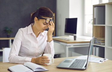 Young tired woman resting during a break from office work, raising her glasses, resting her hand on her face and closing her eyes. Beautiful woman holding a white mug during work break.