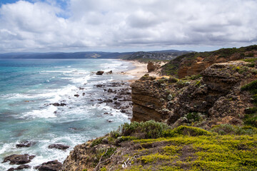 A large panorama view over the rocky coastline of the Australian coast near the Great Ocean Road. Giant waves break on the rocky shore. Steep cliffs break over to the sandy beach. Cloudy sky. 