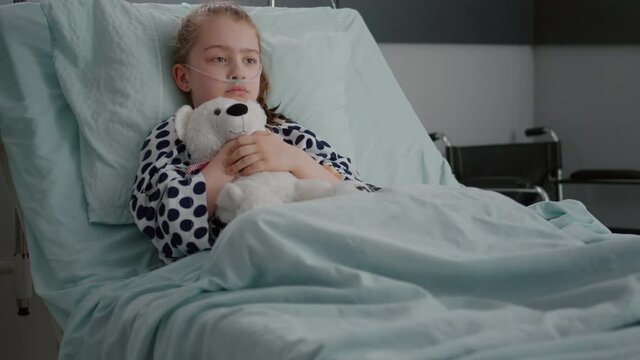 Alone worried child patient wearing oxygen nasal tube resting in bed holding teedy bear in hands recovering disease consultation in hospital ward. Lonely kid suffering fibrosis infection