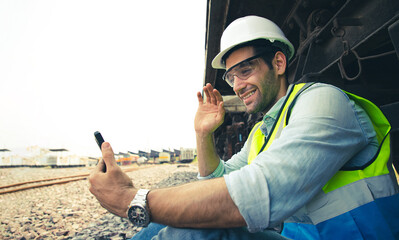 Portrait of handsome engineering using smartphone for video call with  labor with wear hardhat in front of train garage.  Back view of contractor on background of outdoor old train garage.