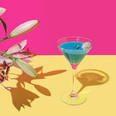 A blue cocktail with a white flower against bright magenta background and warm yellow bottom. Lily flower and its sunny shadow.