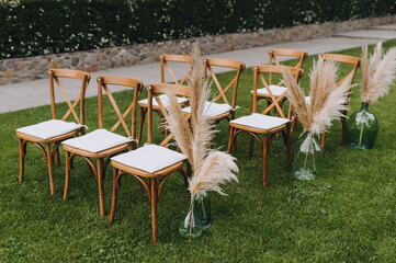 A row of wooden chairs with reed decorations with a vase stands on the green grass in the garden,...