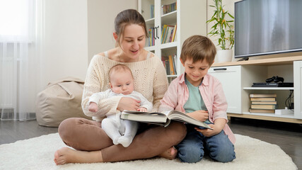 Smiling mother sitting on floor and reading story book to her older and younger sons. Parenting,...