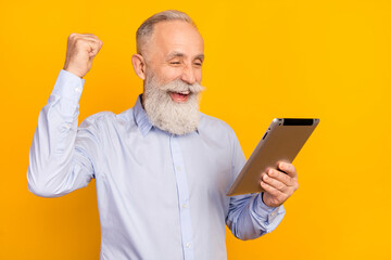 Photo of happy old cheerful man raise fist winner hold tablet celebrate isolated on shine yellow color background