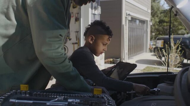 Father guiding son fixing car engine with digital tablet in driveway