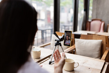 Happy young Caucasian millennial or gen z woman with long brunette hair streaming with smart phone on tripod, shooting social media blog in modern cafe. Influencer using social networks indoor.