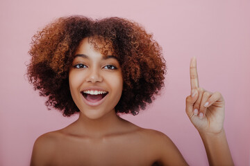 Young beautiful African-American woman with a lush curly hairstyle points her index finger up looks at the camera and smiles on a pink background, a new idea has come, inspiration