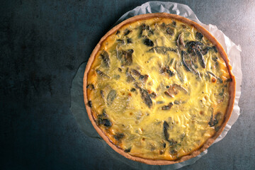 Top view of a freshly baked yellow quiche with mushrooms still on a baking paper, sitting on a dark kitchen table surface.