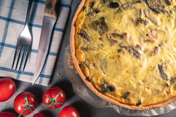 Top view of a freshly baked yellow quiche with mushrooms still on a baking paper, sitting on a dark kitchen table surface with cutlery set, tomatoes, mint leaves.