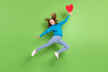 Fototapeta na wymiar Full size photo of happy brunette tailed girl jump fly hair hold red paper heart wear casual clothes isolated on green color background