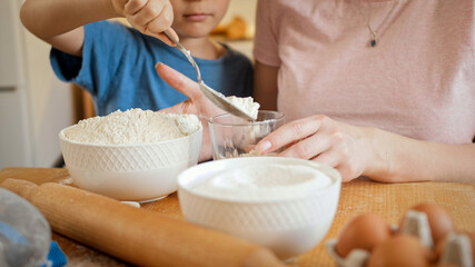 Closeup of little son helping mother pouring flour in bowl for making biscuit or dough. Children cooking with parents, little chef, family having time together, domestic kitchen.