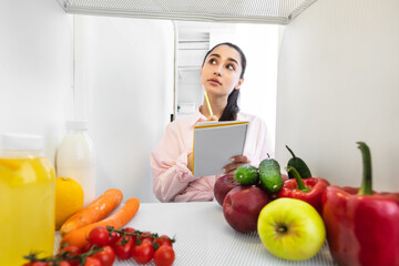 Pensive lady opening fridge and making list of necessary food