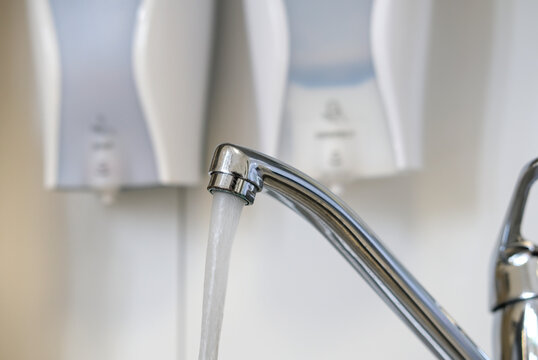 Konzept Hygiene und Sauberkeit: Wasser flie&szlig;t aus einem Wasserhahn und im Hintergrund zwei Spender mit Desinfektionsmittel und Seife an einer wei&szlig;en Wand &ndash;selektiver Fokus 