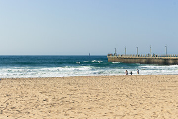 Beach, pier, sailboat, surfers and swimmers. Summer water activities at the coast. Durban, South Africa.